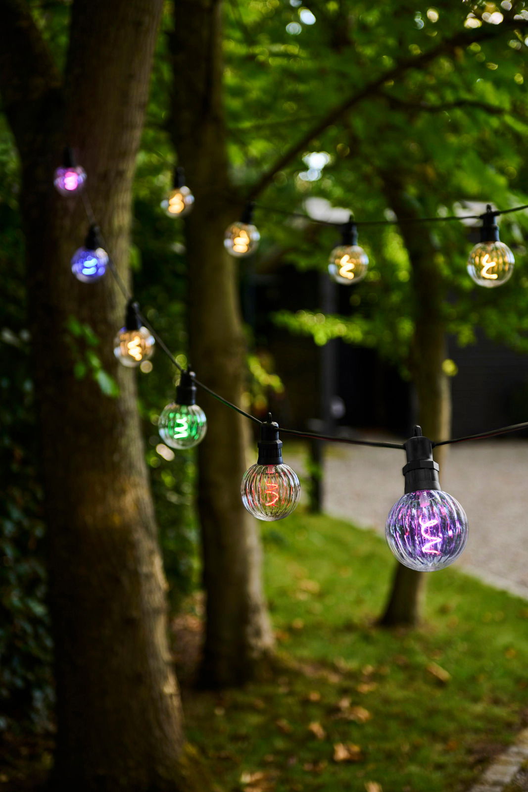 Sirius LED-Lichterkette 10er LUKE Bunte Lichterkette mit 10 LED-Lampen, aufgehängt zwischen Bäumen in einem Garten, aus seitlicher Perspektive fotografiert.