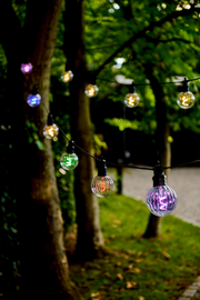 Bunte Lichterkette mit 10 LED-Lampen, aufgehängt zwischen Bäumen in einem Garten, aus seitlicher Perspektive fotografiert.