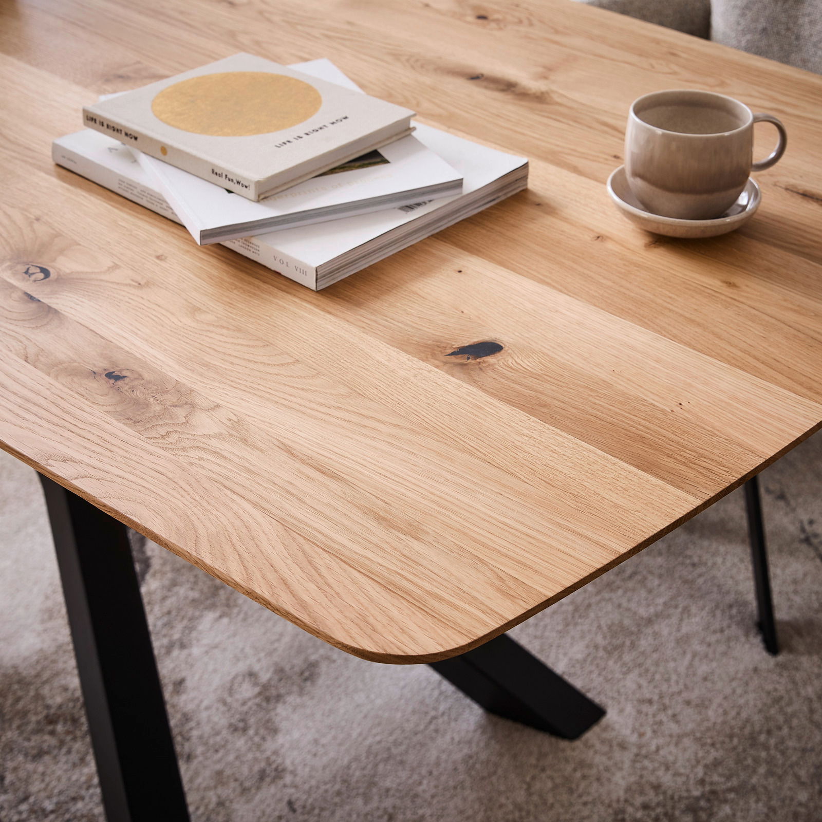 Nahaufnahme eines Esstisches aus Holz mit natürlicher Maserung, darauf liegen Bücher und eine Tasse. Perspektive von schräg oben.