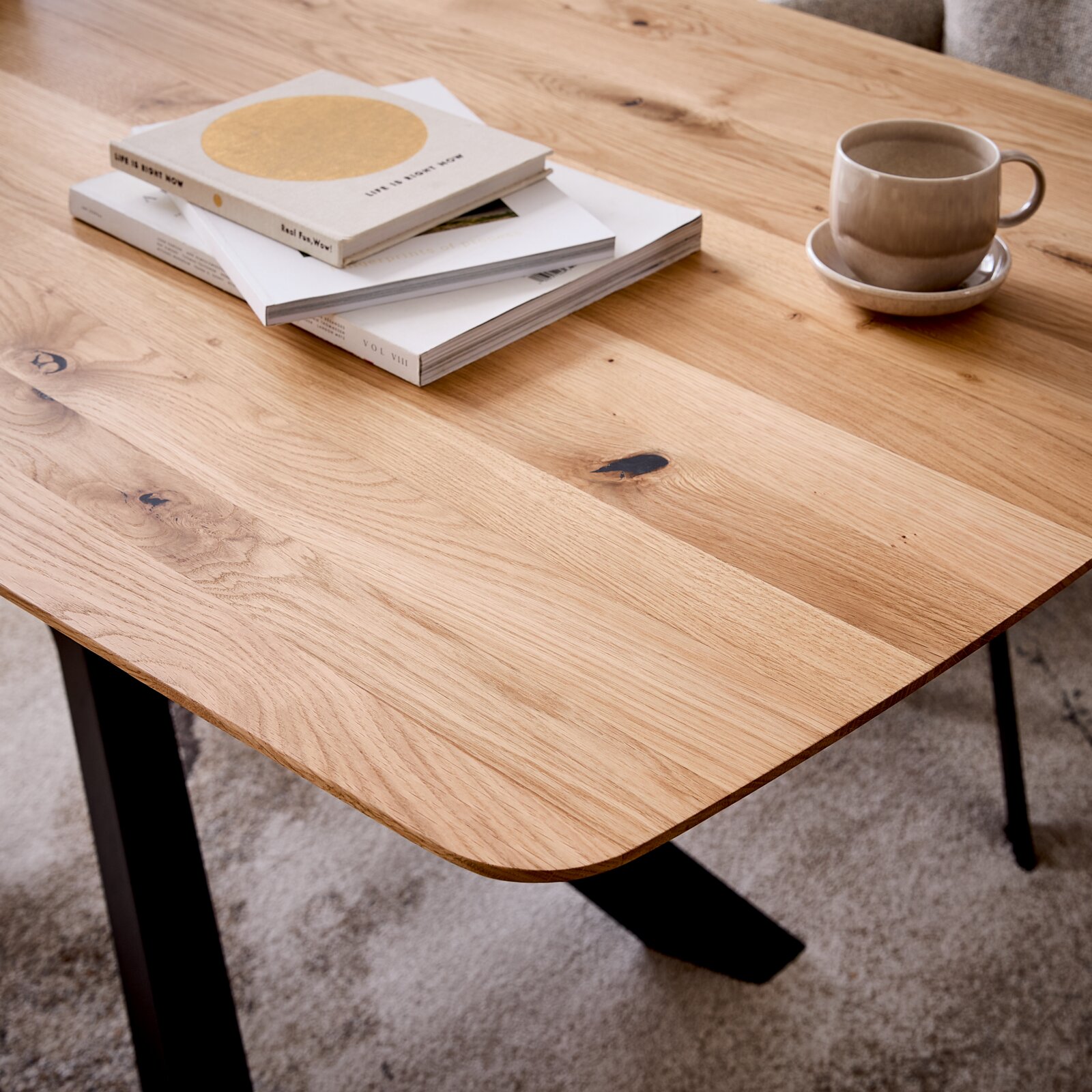Close-up van een houten eettafel met een natuurlijke nerf, met daarop boeken en een kopje. Opname vanuit een schuine hoek van bovenaf.