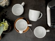 Vier weiße Keramik-Kaffeetassen auf dunkler Arbeitsfläche neben Espressomaschine, Draufsicht von oben