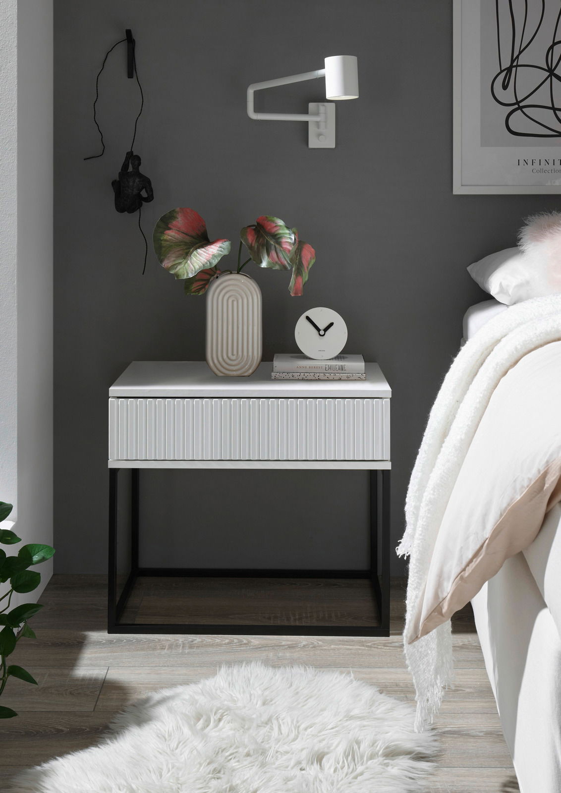 Moderne Nachtkommode in Weiß mit geriffelter Front, schwarzem Metallgestell, dekoriert mit einer Vase und einer Uhr, seitliche Perspektive.