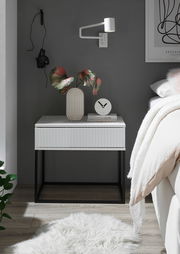 Moderne Nachtkommode in Weiß mit geriffelter Front, schwarzem Metallgestell, dekoriert mit einer Vase und einer Uhr, seitliche Perspektive.