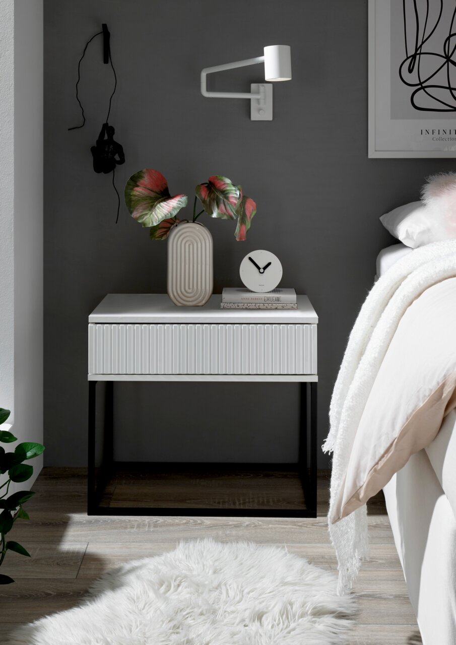 Moderne Nachtkommode in Weiß mit geriffelter Front, schwarzem Metallgestell, dekoriert mit einer Vase und einer Uhr, seitliche Perspektive.