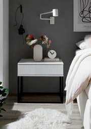 Moderne Nachtkommode in Weiß mit geriffelter Front, schwarzem Metallgestell, dekoriert mit einer Vase und einer Uhr, seitliche Perspektive.