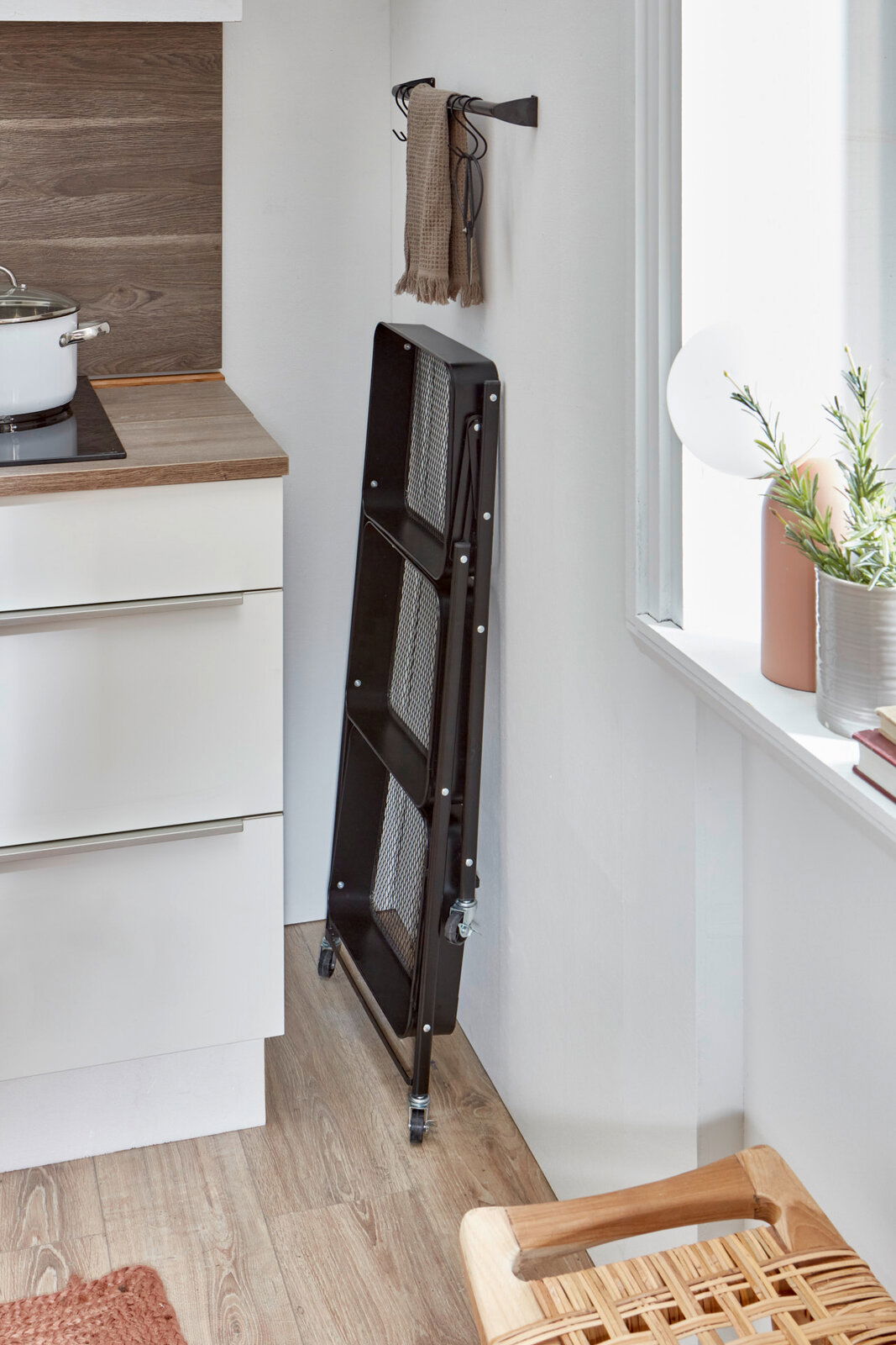 Schwarzer, klappbarer Servierwagen aus Metall mit Gittereinsätzen und Rollen, platzsparend zusammengeklappt an der Wand in einer hellen Küche, seitliche Perspektive.