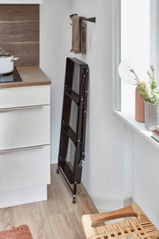 Schwarzer, klappbarer Servierwagen aus Metall mit Gittereinsätzen und Rollen, platzsparend zusammengeklappt an der Wand in einer hellen Küche, seitliche Perspektive.