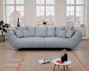 Hellgraues, modernes 3-Sitzer-Sofa mit mehreren Kissen und Metallfüßen im hellen Wohnzimmer vor großen Sprossenfenstern, frontale Ansicht.