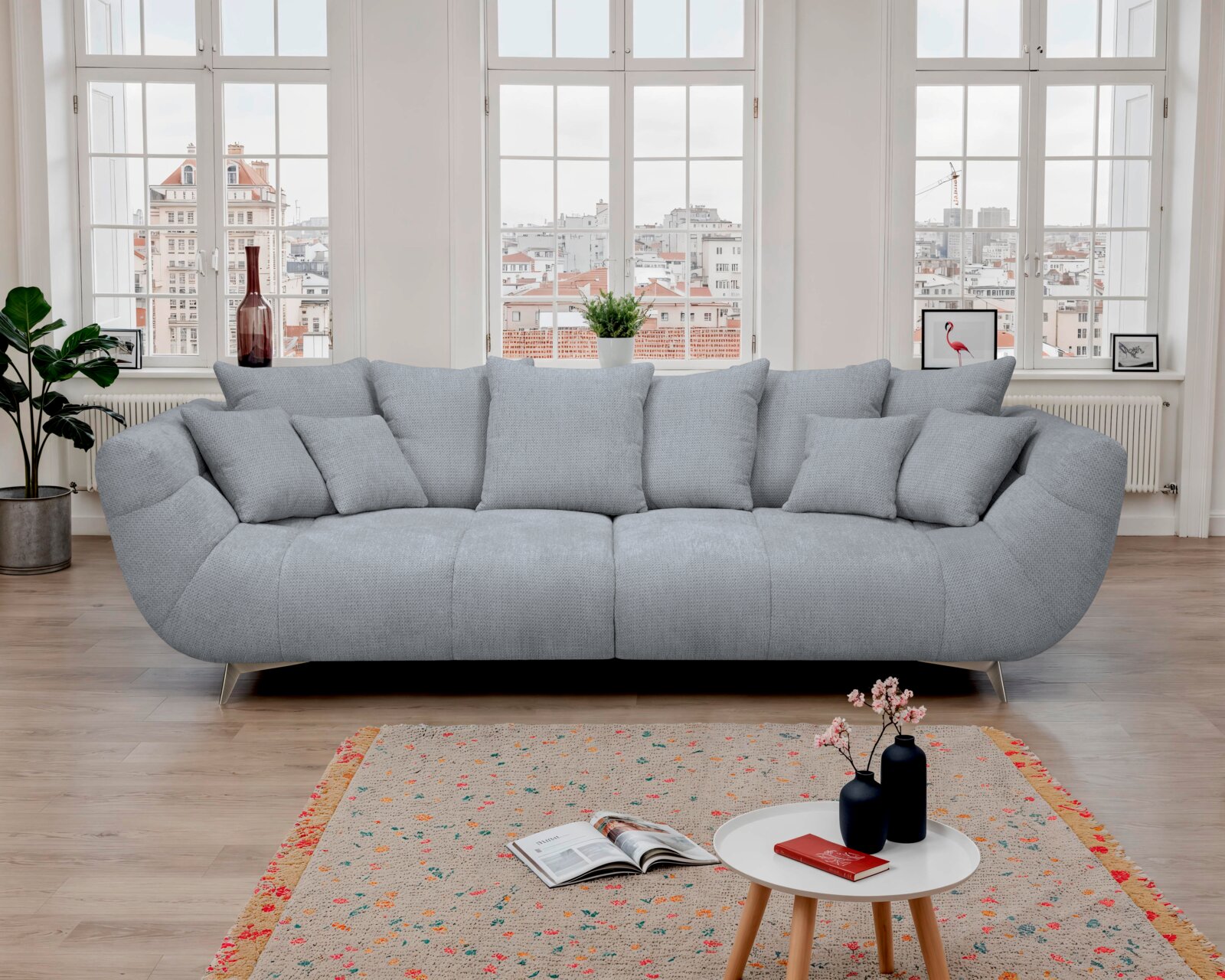 Hellgraues, modernes 3-Sitzer-Sofa mit mehreren Kissen und Metallfüßen im hellen Wohnzimmer vor großen Sprossenfenstern, frontale Ansicht.