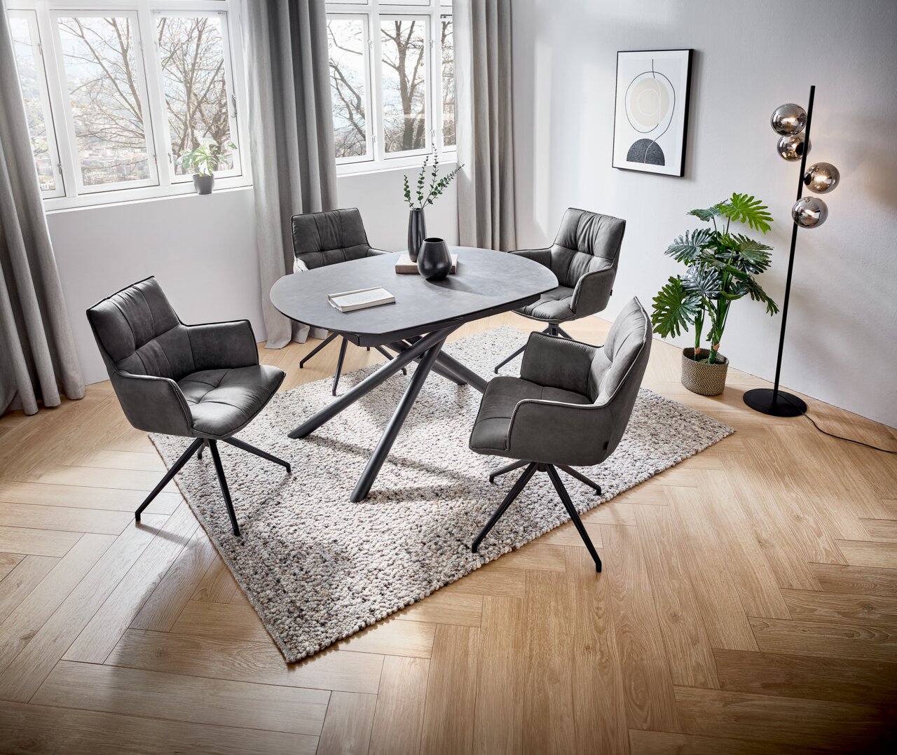 Een moderne eettafel met een ovaal grijs tafelblad en zwarte metalen poten, omringd door vier gestoffeerde stoelen in een lichte ruimte met grote ramen, gezien vanuit vogelperspectief.
