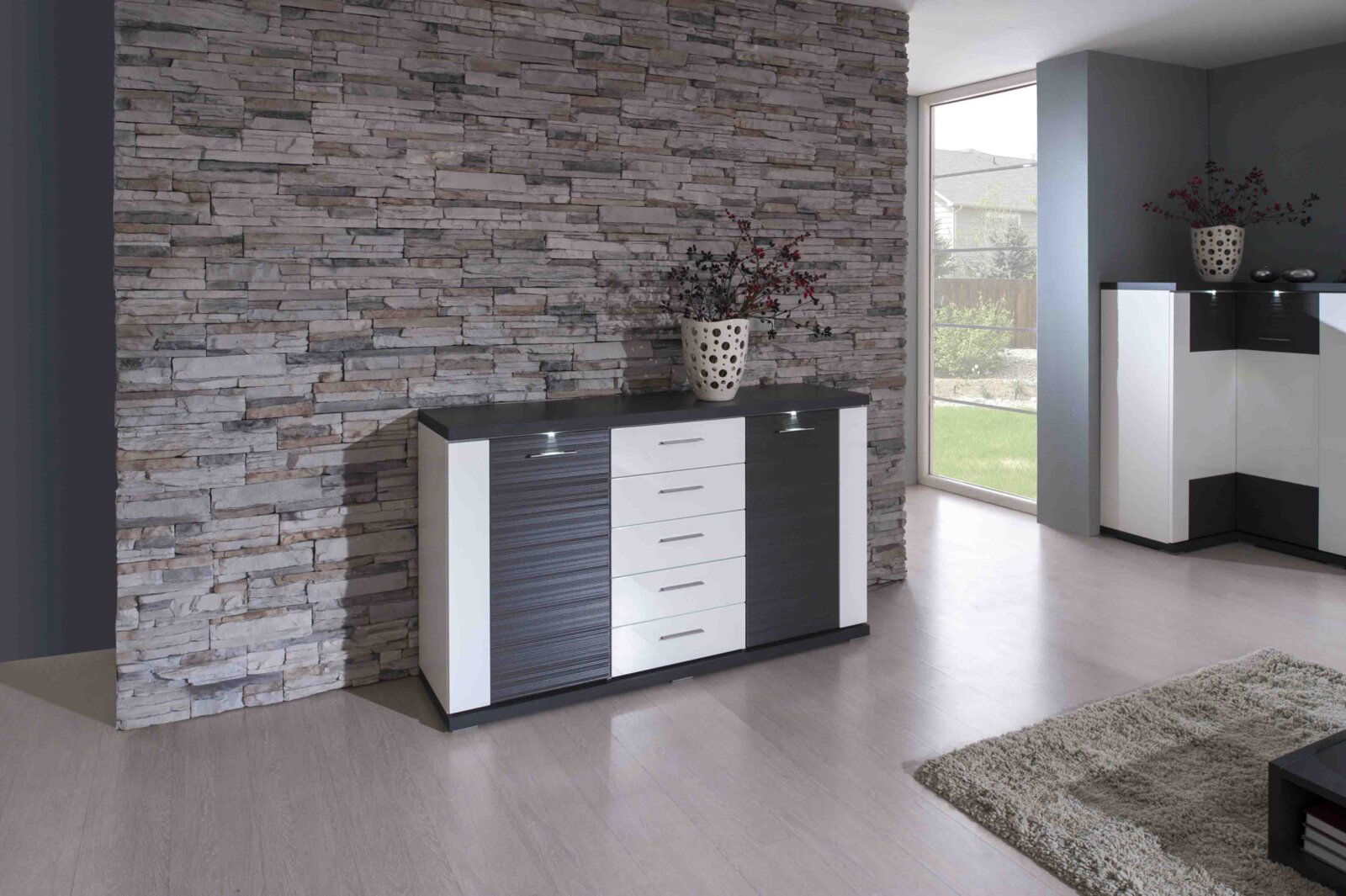 XORA Sideboard MANHATTAN Modernes Sideboard mit dunkler und weißer Front, vor einer Steinwand, aus der Frontalperspektive fotografiert.