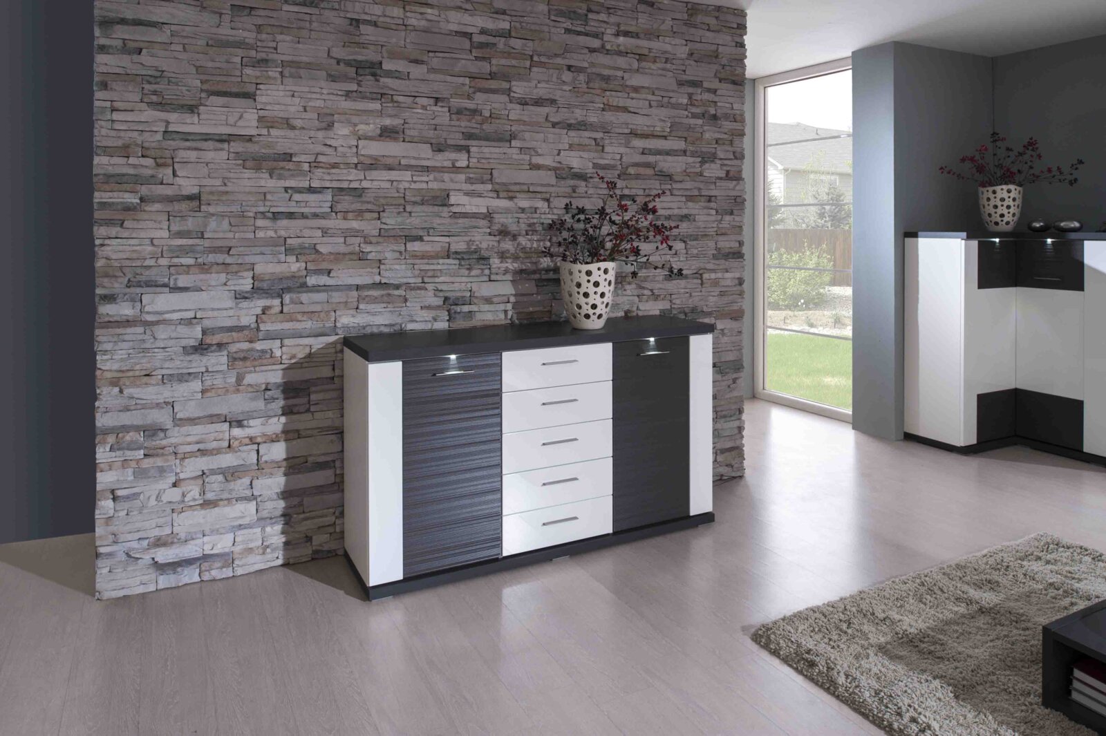 Modernes Sideboard mit dunkler und weißer Front, vor einer Steinwand, aus der Frontalperspektive fotografiert.