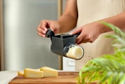 Schwarze Handkurbel-Käsereibe mit Auffangbehälter in Händen, Käse wird gerieben; seitliche Nahaufnahme aus leicht schräger Perspektive über einem Holzbrett mit Käsestücken