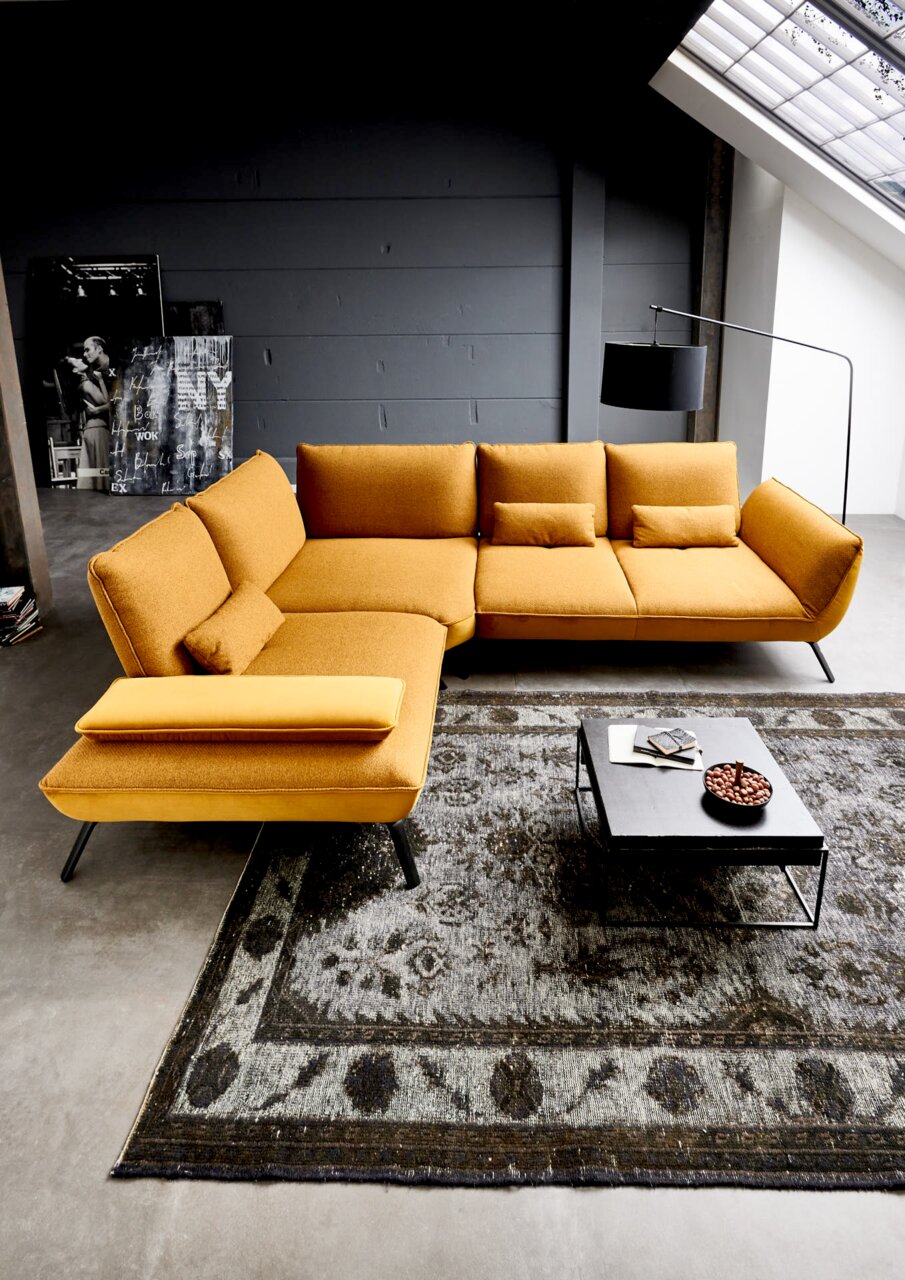 Moderne Eckgarnitur in Senfgelb aus der Vogelperspektive, platziert auf einem gemusterten Teppich neben einem Couchtisch.