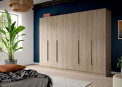 Großer Kleiderschrank in heller Holzoptik mit sechs Türen und langen schwarzen Griffen, in modernem Schlafzimmer vor dunkelblauer Wand, Frontansicht leicht von links