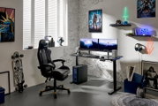 Modernes Gaming-Setup im Zimmer mit schwarzem Gaming-Stuhl, höhenverstellbarem Schreibtisch, Dual-Monitoren und PC-Tower mit blauer Beleuchtung, seitliche Perspektive von links vorn
