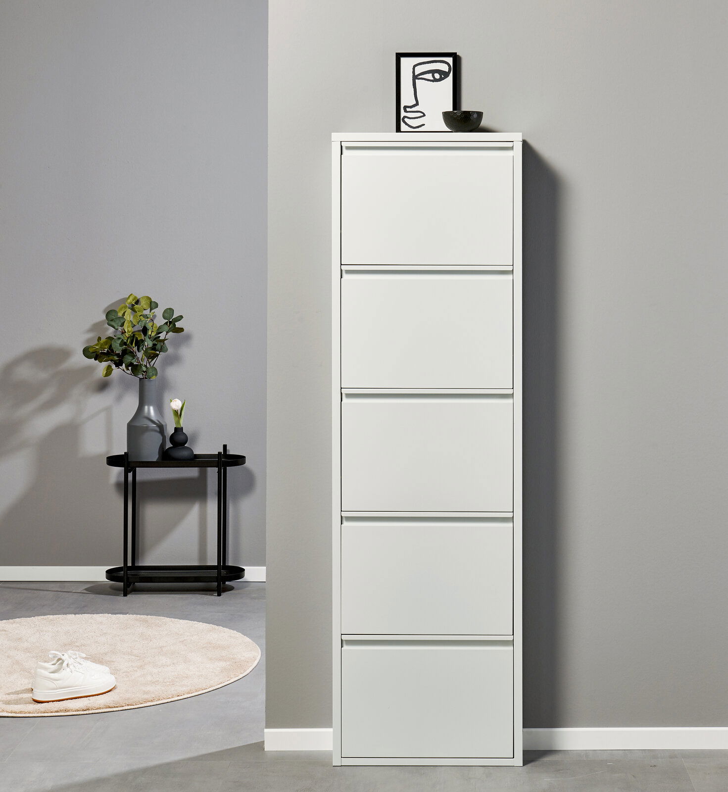 Schlanker, weißer Schuhschrank mit fünf Klappfächern vor grauer Wand, frontal aus Augenhöhe fotografiert; daneben ein schwarzer Beistelltisch mit Vase und Deko.