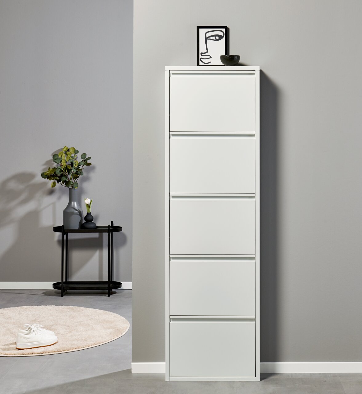 Een slanke, witte schoenenkast met vijf klapdeurtjes tegen een grijze muur, van voren gefotografeerd vanaf ooghoogte; ernaast een zwart bijzettafeltje met een vaas en decoratie.