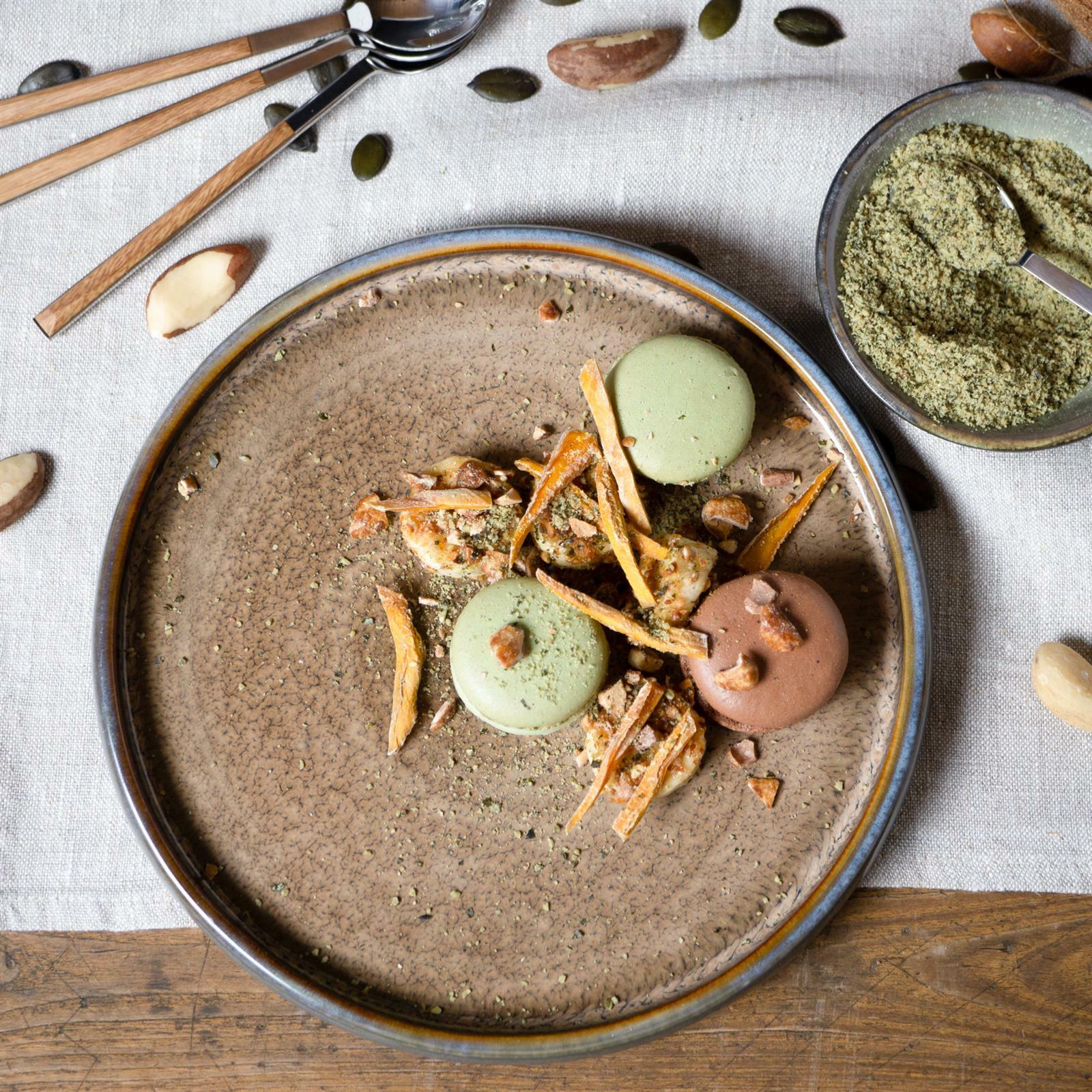 Vogelperspektive auf einen rustikalen, beige-braunen Teller mit dekorativen Macarons und Nüssen, umgeben von Besteck und Schalen auf einem Leinentuch.