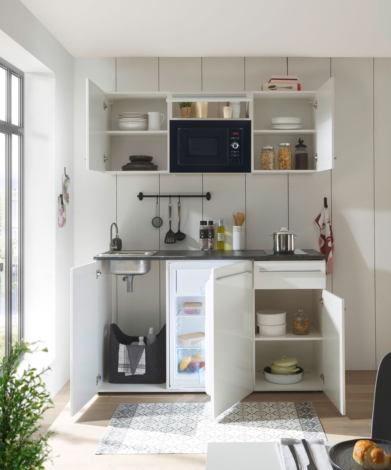 Moderne Single-Küche mit offenem Schrank, Mikrowelle, Spüle und Kühlschrank, aus der Frontalperspektive fotografiert.