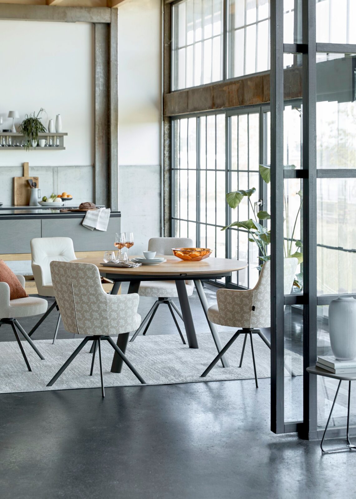 JOOP! Esstisch STUDIO J62 Schräg von vorn fotografierter moderner Essbereich mit rundem Holzesstisch und gepolsterten Stühlen auf Teppich, vor großer Fensterfront in heller Loftküche