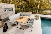 Modernes Outdoor-Ess-Set auf einer Poolterrasse: hellgraue gepolsterte Eckbank mit zwei passenden Stühlen und rechteckigem Holztisch mit schwarzem Metallgestell, fotografiert in schräger Perspektive von vorne oben.