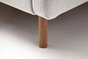 Close-up van een gestoffeerd bed met een lichtgrijze stoffen bekleding en een houten poot, gezien vanaf de zijkant.