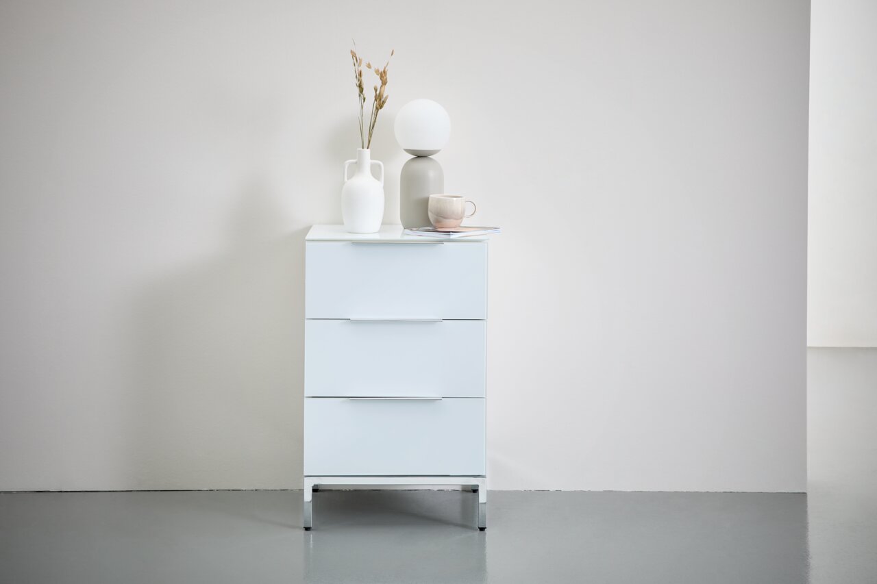 Weißer Schrank mit drei Schubladen auf Metalluntergestell, frontal fotografiert. Dekoriert mit Vasen und einer Tasse auf der Oberseite.