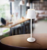Weiße Akku-Tischleuchte VANNIE auf einem Tisch in einem modernen Café, seitliche Perspektive.