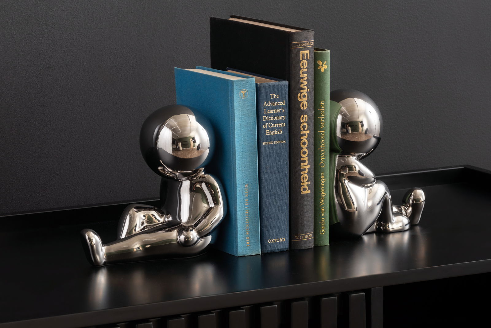 Zwei glänzende, silberne Buchstützen in Form von stilisierten Figuren, die auf einem Regal sitzen und Bücher zwischen sich halten. Perspektive von vorne.