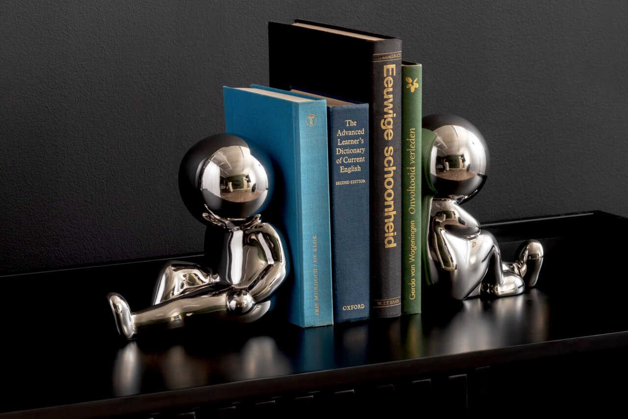 present time Buchhalter 2-tlg. JOEY Zwei glänzende, silberne Buchstützen in Form von stilisierten Figuren, die auf einem Regal sitzen und Bücher zwischen sich halten. Perspektive von vorne.