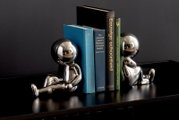 Twee glanzende, zilveren boekensteunen in de vorm van gestileerde figuurtjes die op een plank zitten en boeken tussen zich in houden. Vooraanzicht.
