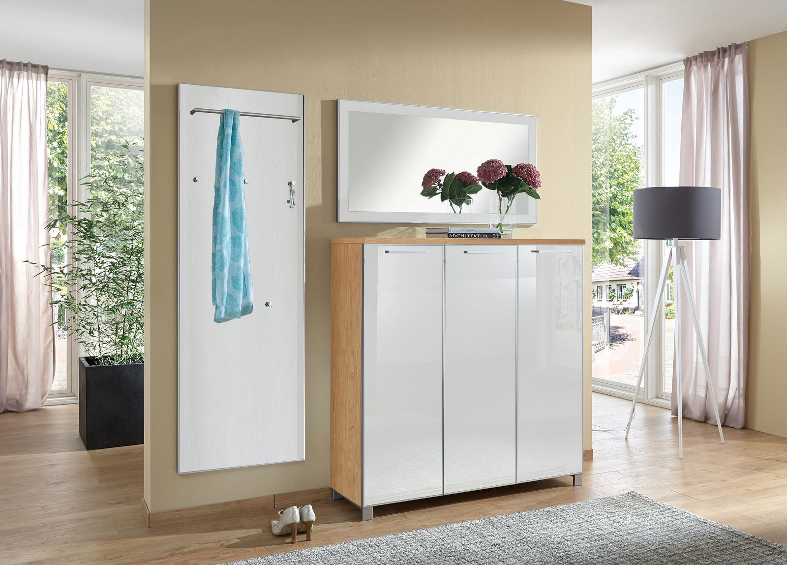 Garderoben-Set bestehend aus einem weißen Wandpaneel mit Kleiderstange und Haken, einem großen Spiegel und einem weißen Schrank mit Holzrahmen, in einem modernen, hellen Raum mit großen Fenstern, aus frontaler Perspektive.