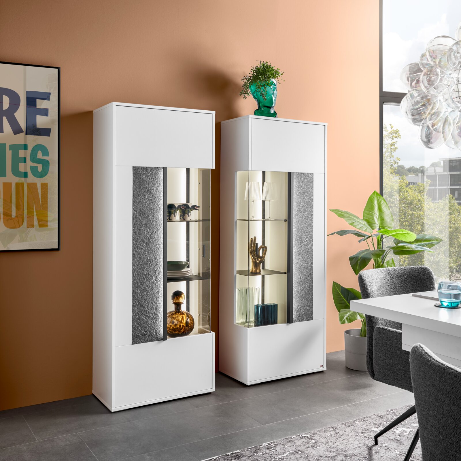 Set one by Musterring Highboard/Vitrine links DELTONA Modernes Highboard mit Glasvitrine, links im Bild, in einem stilvollen Wohnzimmer mit Pflanzen und Kunst an der Wand, seitliche Perspektive.