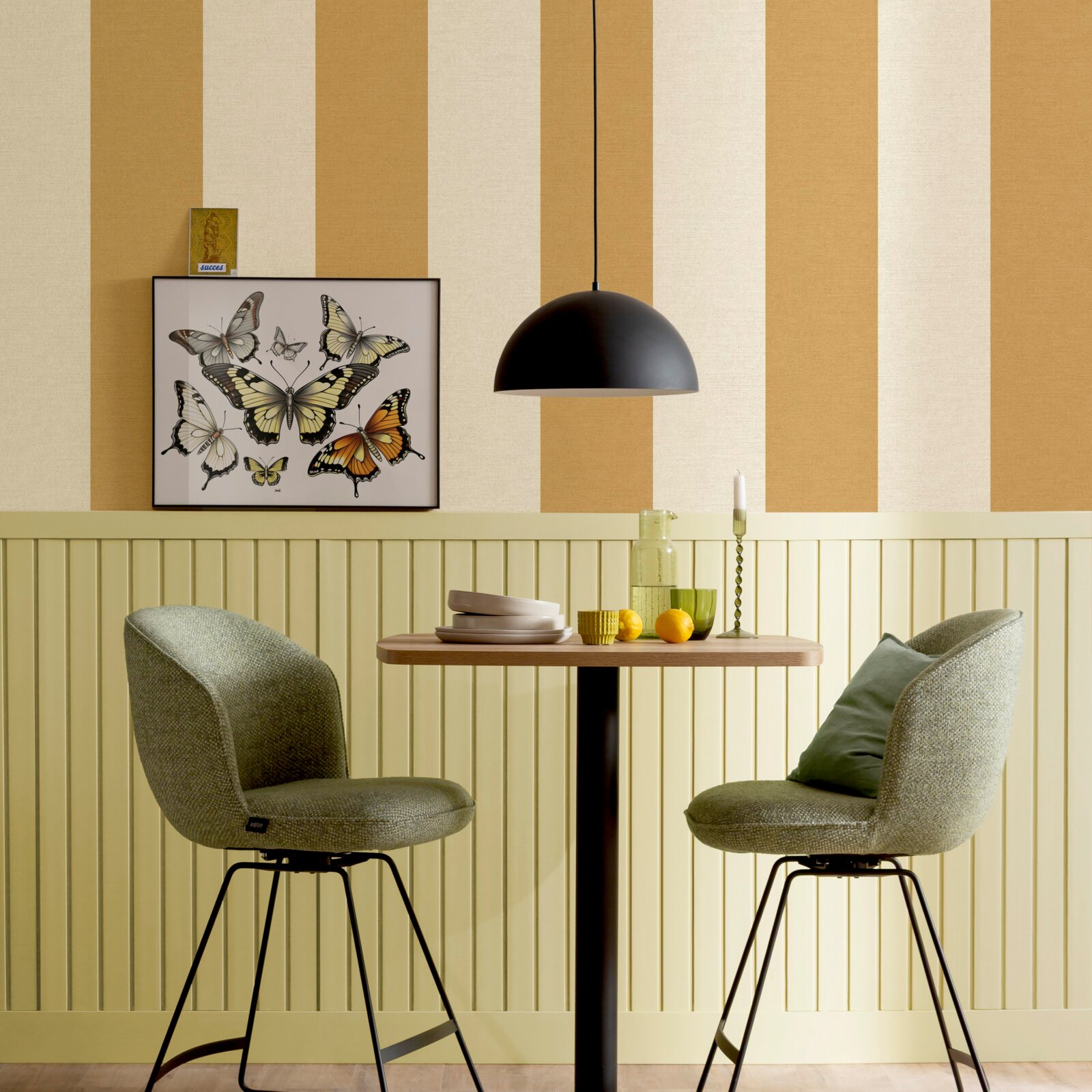Schwarze, halbrunde Pendelleuchte über einem kleinen Holztisch mit zwei grünen Polsterstühlen vor einer gestreiften Wand und Schmetterlingsbild, Frontansicht auf Augenhöhe.