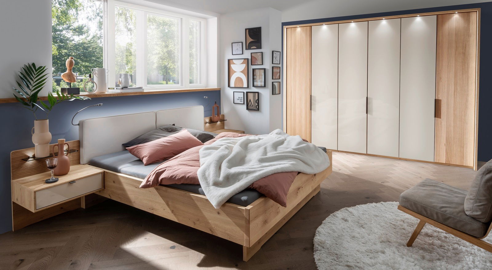 Modernes Schlafzimmer mit einem Nachtschrank aus Holz, links neben einem Bett mit rosa und grauer Bettwäsche, im Tageslicht von einem großen Fenster.