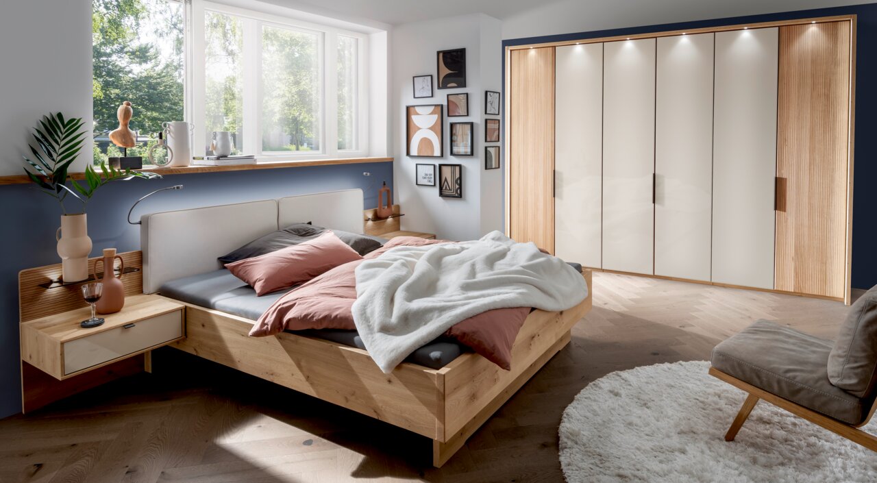 Modernes Schlafzimmer mit einem Nachtschrank aus Holz, links neben einem Bett mit rosa und grauer Bettwäsche, im Tageslicht von einem großen Fenster.