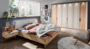 Modernes Schlafzimmer mit einem Nachtschrank aus Holz, links neben einem Bett mit rosa und grauer Bettwäsche, im Tageslicht von einem großen Fenster.