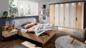 Een moderne slaapkamer met een houten nachtkastje, links naast een bed met roze en grijs beddengoed, verlicht door het daglicht dat door een groot raam naar binnen valt.