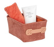 Rechteckiger Badkorb Anela mini in einem warmen Braunton, gefüllt mit einem orangefarbenen Handtuch und einer weißen Tube. Der Korb hat eine dekorative Lasche an der Seite. Perspektive: leicht von oben.