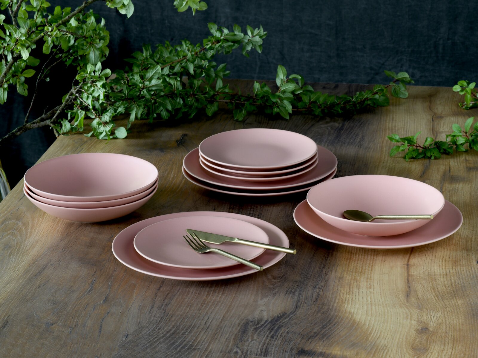Creatable Tellerset 12-tlg SOFT TOUCH Pastellrosa Geschirr-Set mit Tellern und Schalen auf rustikalem Holztisch, dekoriert mit grünen Zweigen; Perspektive: leicht erhöhte, schräge Vorderansicht.