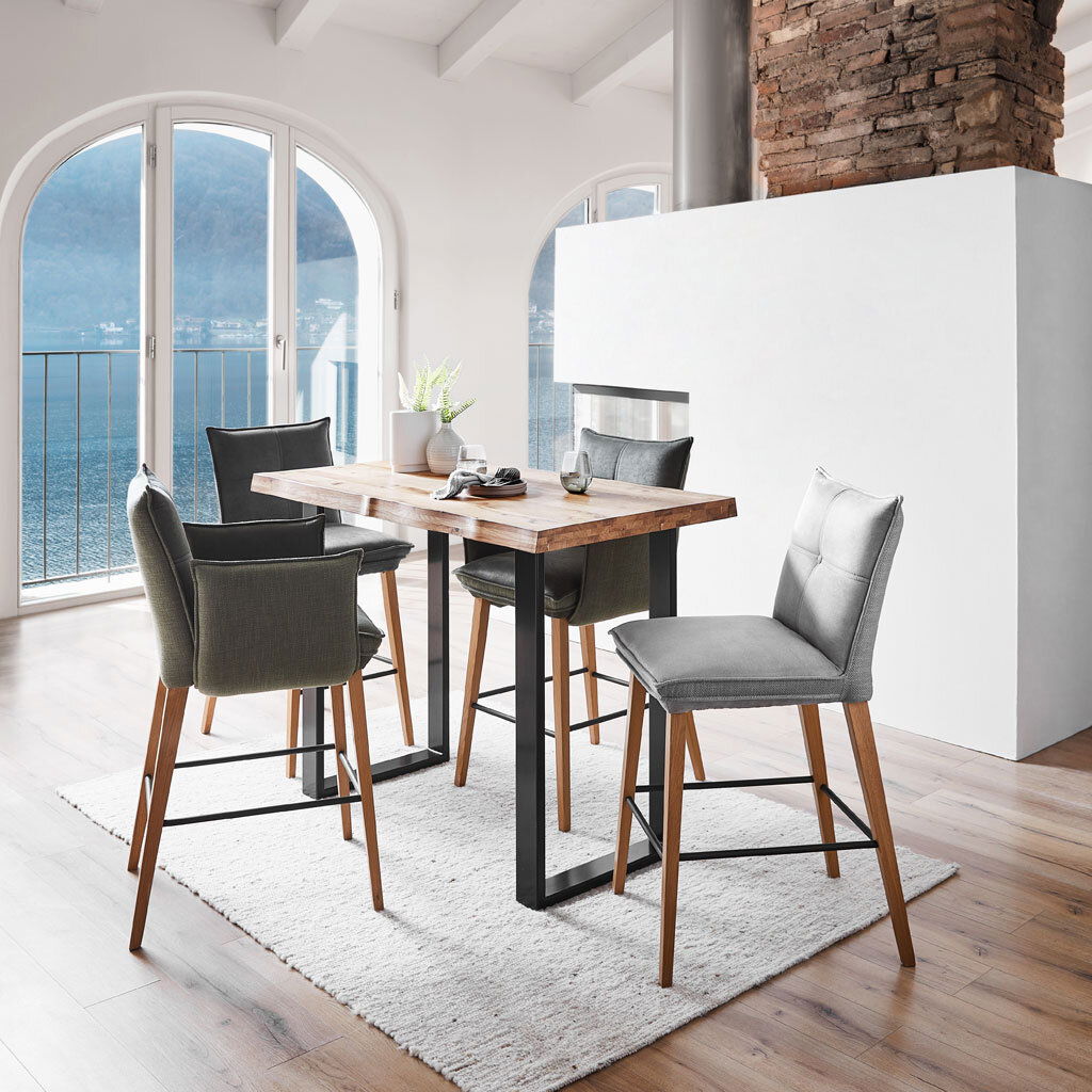 Moderner Bartisch mit Holzplatte und schwarzen Metallbeinen, umgeben von stilvollen Barhockern in einem hellen Raum mit Blick auf einen See. Perspektive: seitlich.