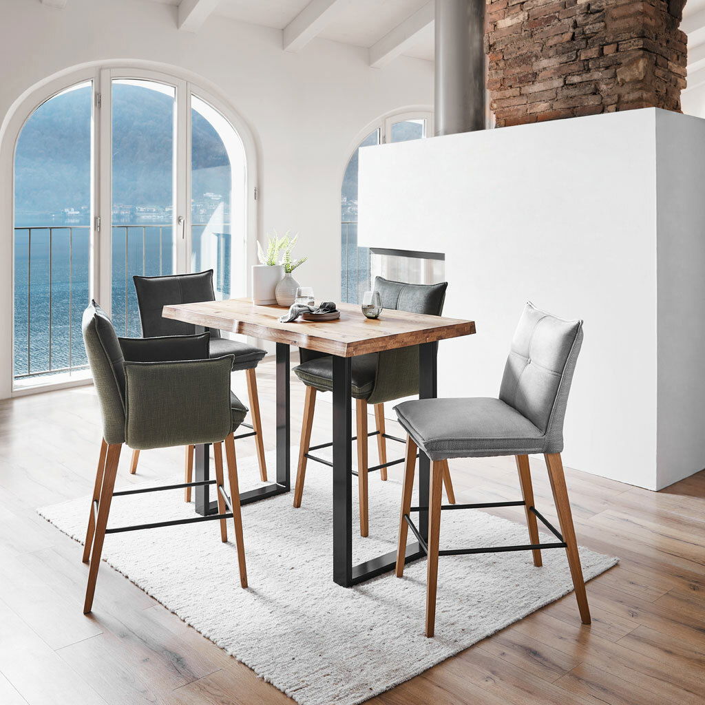 Moderner Bartisch mit Holzplatte und schwarzen Metallbeinen, umgeben von stilvollen Barhockern in einem hellen Raum mit Blick auf einen See. Perspektive: seitlich.