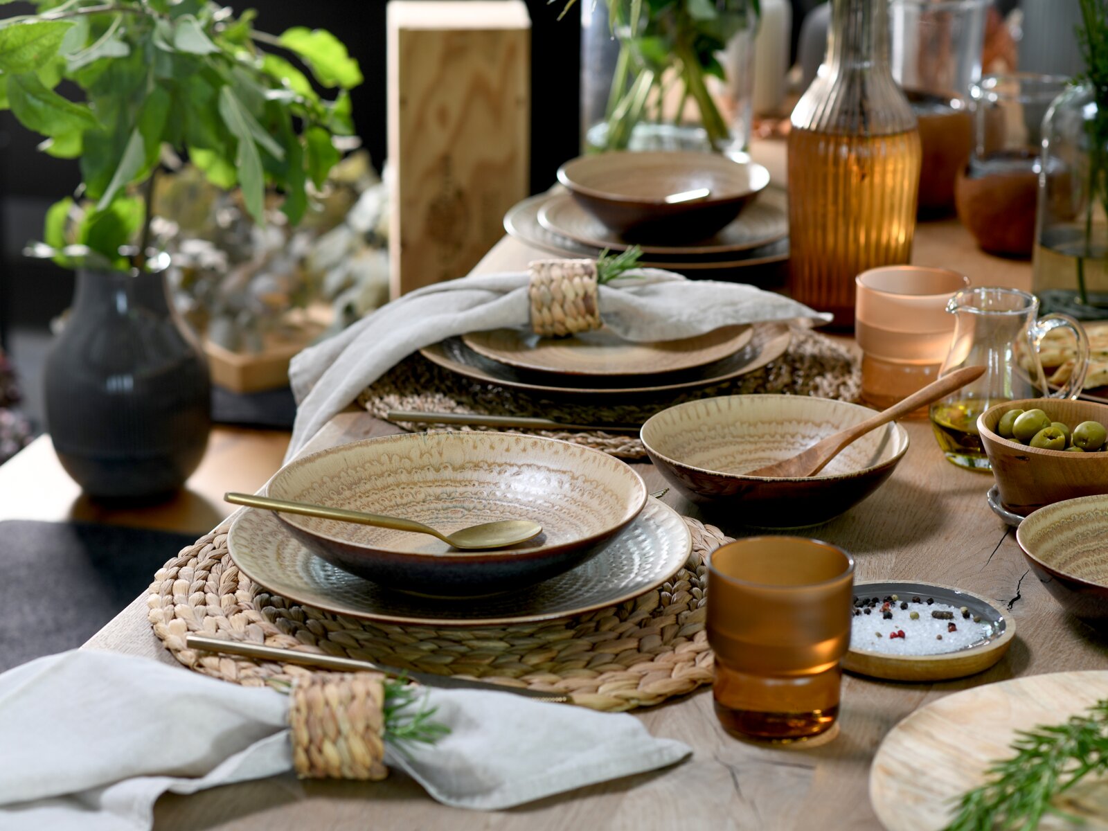 Rustikales Tischgedeck mit gemusterten Keramikschalen und Tellern auf geflochtenen Platzsets, goldfarbenem Besteck, amberfarbenen Gläsern, Leinservietten mit Serviettenringen sowie Oliven und Öl auf Holztisch, perspektivisch aus leicht schräger Seitenansi