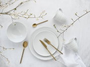 Flatlay aus der Vogelperspektive: weißes Porzellan-Geschirrset mit strukturiertem Rand (Teller und Schale), zwei weißen Tassen, goldfarbenem Besteck und zarten Blütenzweigen auf hellem Untergrund