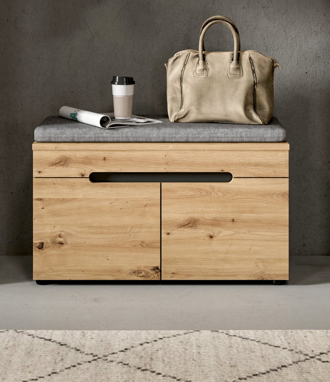 Frontale Ansicht einer modernen Holz-Sitzbank mit grauem Sitzkissen und zwei Türen, darauf eine beige Handtasche, ein Coffee-to-go-Becher und eine Zeitung vor grauer Wand.