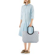 Frau in hellblauem Kleid hält eine graue, gesteppte Tasche mit großen, schwarzen Griffen. Die Tasche hat ein Rhombus-Muster und wird von der Seite gezeigt.