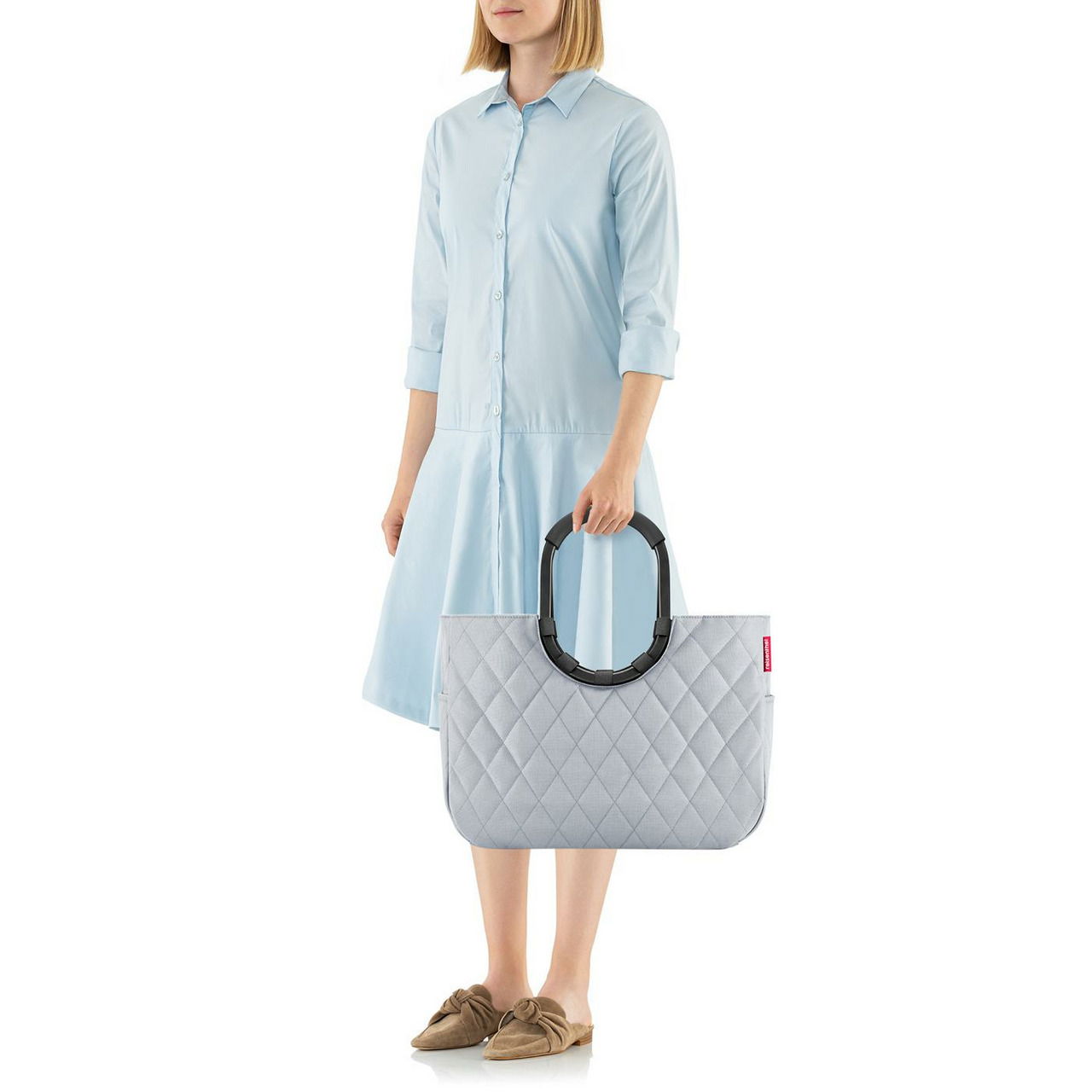 Frau in hellblauem Kleid hält eine graue, gesteppte Tasche mit großen, schwarzen Griffen. Die Tasche hat ein Rhombus-Muster und wird von der Seite gezeigt.