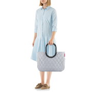 Frau in hellblauem Kleid hält eine graue, gesteppte Tasche mit großen, schwarzen Griffen. Die Tasche hat ein Rhombus-Muster und wird von der Seite gezeigt.