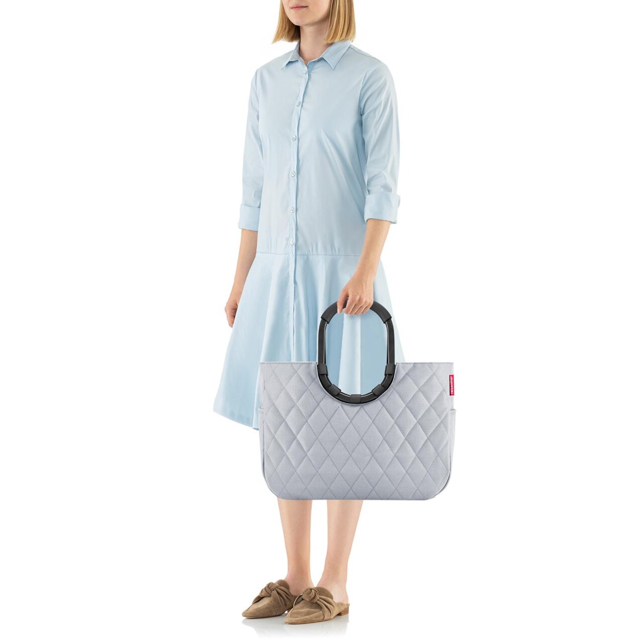 Frau in hellblauem Kleid hält eine graue, gesteppte Tasche mit großen, schwarzen Griffen. Die Tasche hat ein Rhombus-Muster und wird von der Seite gezeigt.